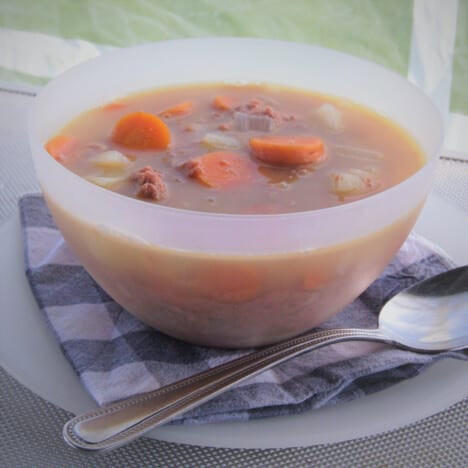 A white camp bowl of corned beef stew ready to eat on a blue checkered napkin.
