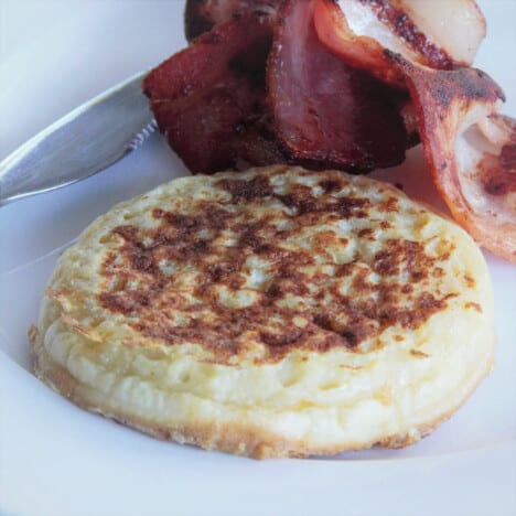 A single, cooked crumpet French toast, golden brown on top, on a plate with fried bacon slices.