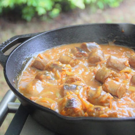 Sliced sausages, chopped onions, and grated carrots simmering in the curry sauce mixture in a cast iron frying pan.