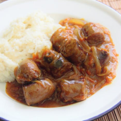 Completed curried sausages ready to serve on a white plate with a side of mashed potatoes.