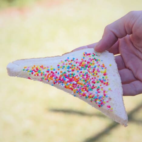 A hand holding a triangular slice of white bread spread with margarine and topped by multi-colored sprinkles.