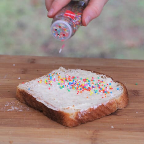 Sprinkles being added to a piece of buttered white bread on a wooden cutting board.