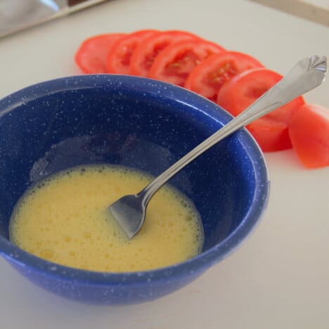 A blue camp bowl with an egg mixture it it and sliced tomatoes behind.