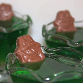 Close up of a clear glass cup with fluted edge filled with set green jelly, a chocolate frog partially submerged at the rim.