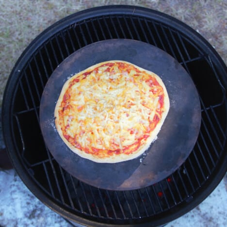 Overhead view of an almost fully cooked ham and pineapple pizza on a pizza stone that is sitting on a grill in a BBQ.