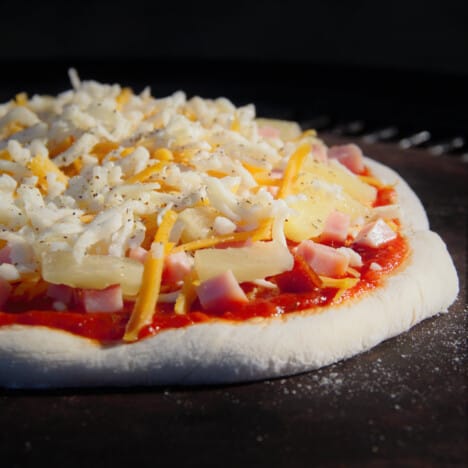 Close up of an assembled ham and pineapple pizza resting on a pizza stone, ready to be cooked.