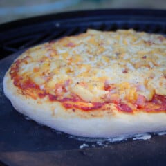 Close up of an almost fully cooked ham and pineapple pizza on a pizza stone that is sitting on a grill in a BBQ.