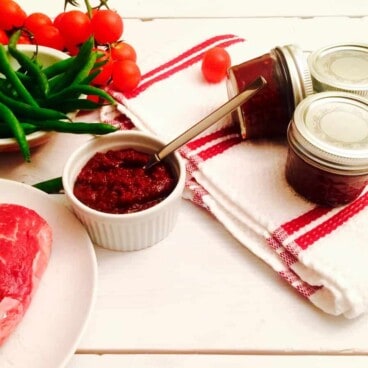 A ramekin of harissa ready to use, sitting next to a raw beef steak, green beans, and three small jars of canned harissa.