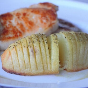 Baked Hasselback potato served as a side on a white plate along with fried pork steaks.