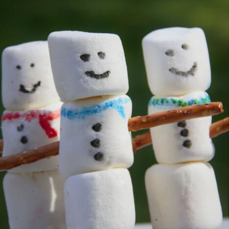 Close up of three marshmallow snowmen, each with a painted face, scarf, and buttons on their body along with pretzel arms.