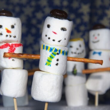 Four marshmallow snowmen on skewers, each decorated with a painted face and scarf, pretzel arms, and a candy for a hat.