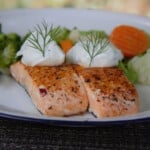Close up of two small cooked salmon fillets, each filet topped with a dollop of yogurt dill sauce and garnished by a dill sprig.