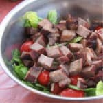 A large, metal bowl containing barbecued steak pieces on a salad of sliced lettuce, cherry tomatoes and shredded cheese.