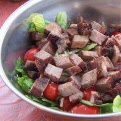A large, metal bowl containing barbecued steak pieces on a salad of sliced lettuce, cherry tomatoes and shredded cheese.
