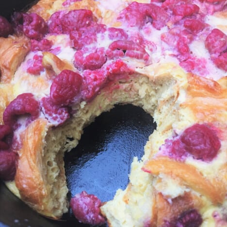 Close up of fully cooked, golden brown pudding studded with raspberries in a Dutch oven, with one scoop removed.