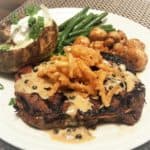 A grilled steak covered in sauce topped with crispy onion straws on a plate with side dishes, ready to eat.