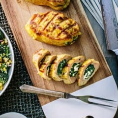 Grilled pork chops resting on a wood cutting board, with one chop sliced into sections to show the spinach and feta stuffing.