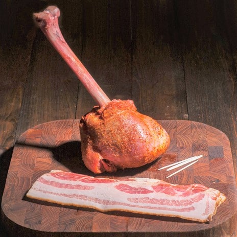A raw lollipop style turkey drumstick on a chopping board with bacon and toothpicks.