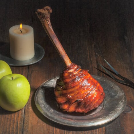A fully cooked and glazed turkey drumstick served on a silver plate next to a candle and fresh apples.