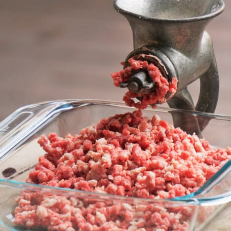 An old hand-cranked grinder with coarsely ground beef sitting in a glass dish below.
