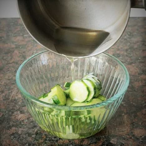 A glass bowl filled with sliced cucumbers with a clear liquid being pour into it from a saucepan.