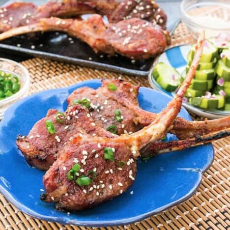Three grilled lamb chops are decratively served on a blue place with a side of pickled cucumber.