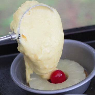 An ice cream scoop placing cake mix in a small cake tin on top of a pineapple ring with a cherry in it.