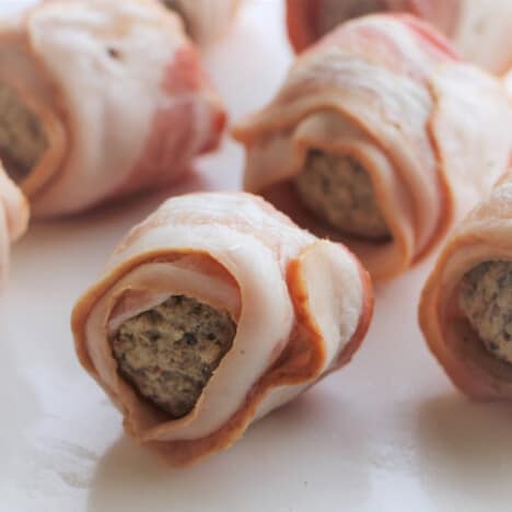 Raw meatballs wrapped with bacon sitting on a white chopping board.