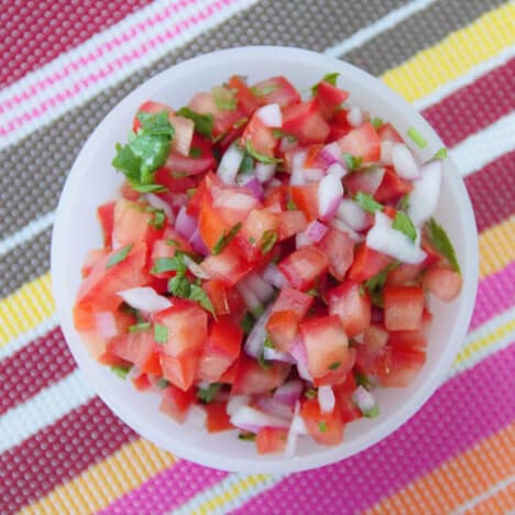 Looking down into a white bowl with pico de gallo on a colorful striped tablecloth.