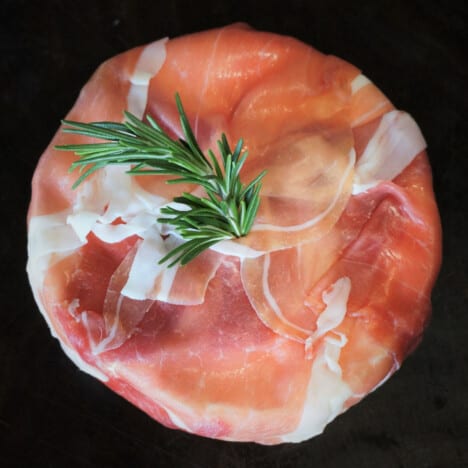 Looking down on a round of Brie wrapped in prosciutto with a sprig of rosemary.