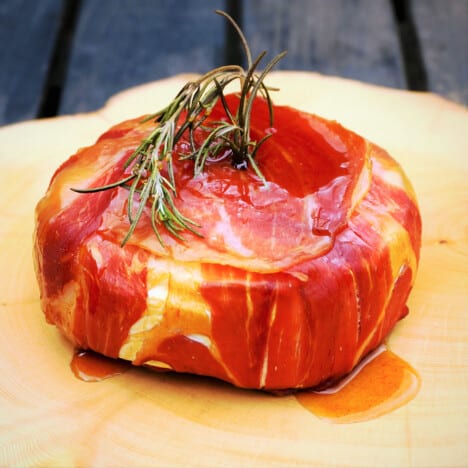 A smoked prosciutto-wrapped Brie is on a chopping board waiting to be served.
