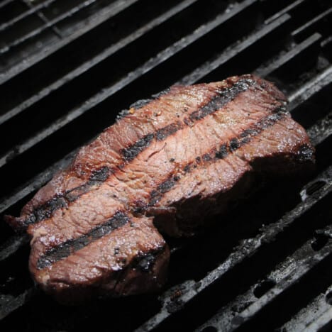 A single lone steak sitting on grill grates having already been cooked on one side.