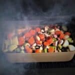 Chopped vegetables and wine covering Lamb shanks in an uncovered baking dish sitting in a smoker, ready to cook.
