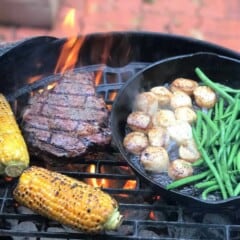 Steak, corn on the cob, and a cast iron skillet containing green beans and scallops, all cooking on a grill over hot coals.
