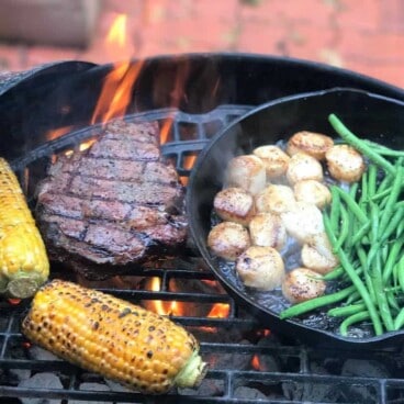 Steak, corn on the cob, and a cast iron skillet containing green beans and scallops, all cooking on a grill over hot coals.