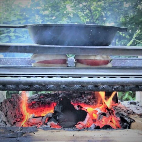 A skillet is pressing down a sheet pan on top of two caprese sandwiches on a grill over coals.