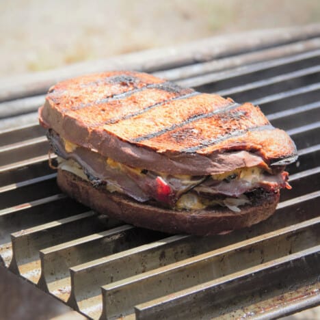 A whole reuben sandwich sits on the grill with dark hatch marks.