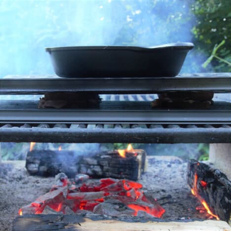 A cast iron pan sits on a baking sheet on two sandwiches on a grill over coals.