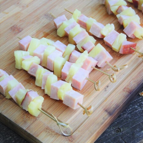 Uncooked ham and pineapple kebabs laid out on a wooden cutting board.