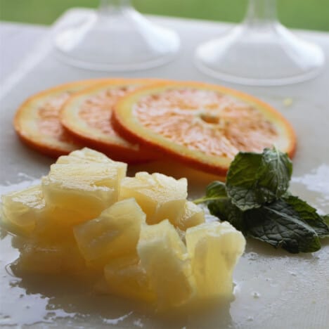 A white chopping board with pineapple chunks, orange slices, and mint leaves on it.