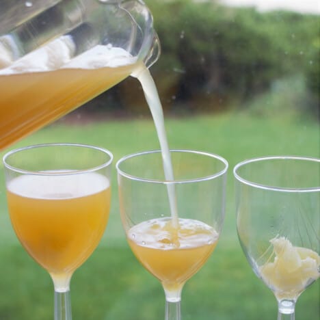A pitcher of pineapple orange mocktail being poured into a glass with pineapple in the base with a full one to the left and empty to the right.