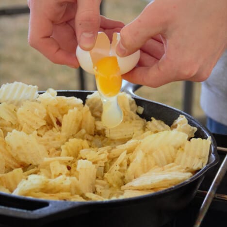 An egg being cracked into a prepared potato chip hash in a skillet.