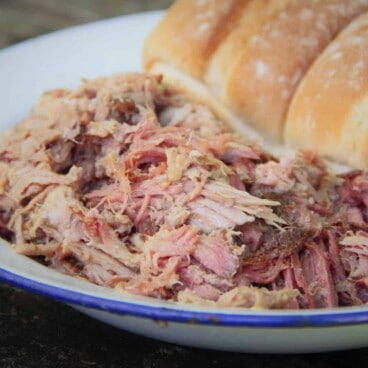 Shredded smoked pork on a white plate with buns in the background.