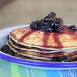 A serving of stacked ricotta pancakes topped with blueberries on a white camp plate.