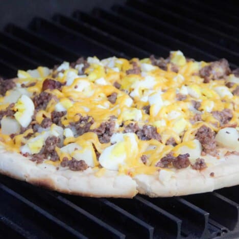 Looking into a breakfast pizza with hard-boiled eggs, cheese, and sausage, on a grill grate.