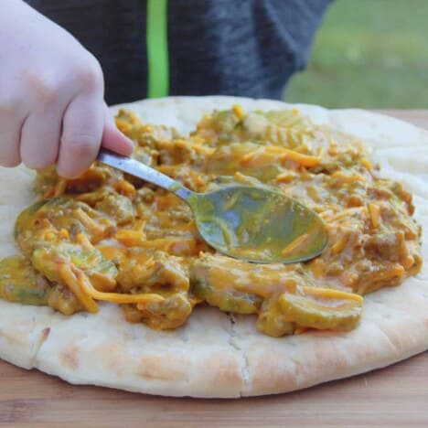 A hand holding a spoon spreads the cheeseburger base on a cooked pizza dough.