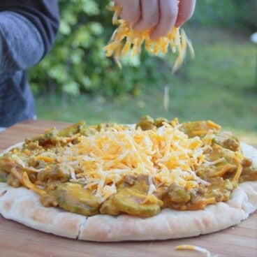 A cheeseburger pizza sits on a cutting board, with grass in the background, while a hand sprinkles cheese on top.