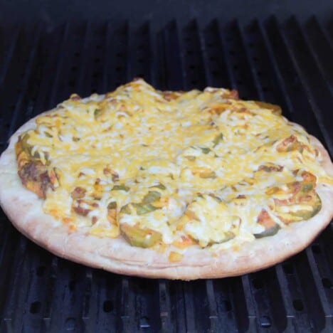 A large cheeseburger pizza cooks on a grill.