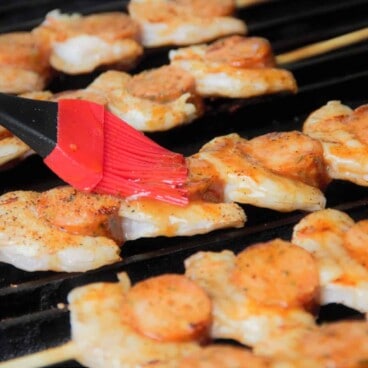 Looking across shrimp and smoked sausage skewers on a grill while they are being glazed with a red brush.