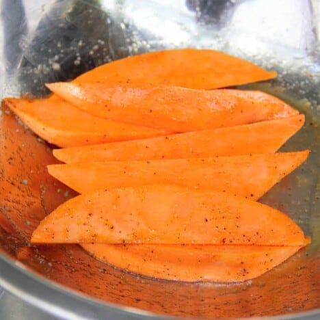 A stainless steel bowl holds a piled of raw sweet potato wedges tossed in oil, salt, and pepper.